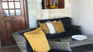 Un canapé en osier agrémenté de coussins jaune moutarde et blancs, une table avec un plateau en verre et une porte en bois dotée d’une fenêtre.
