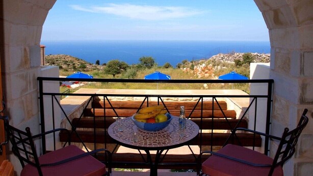 Un balcon orné d’une arche en pierre, une table avec un saladier rempli de fruits et une vue sur la mer.
