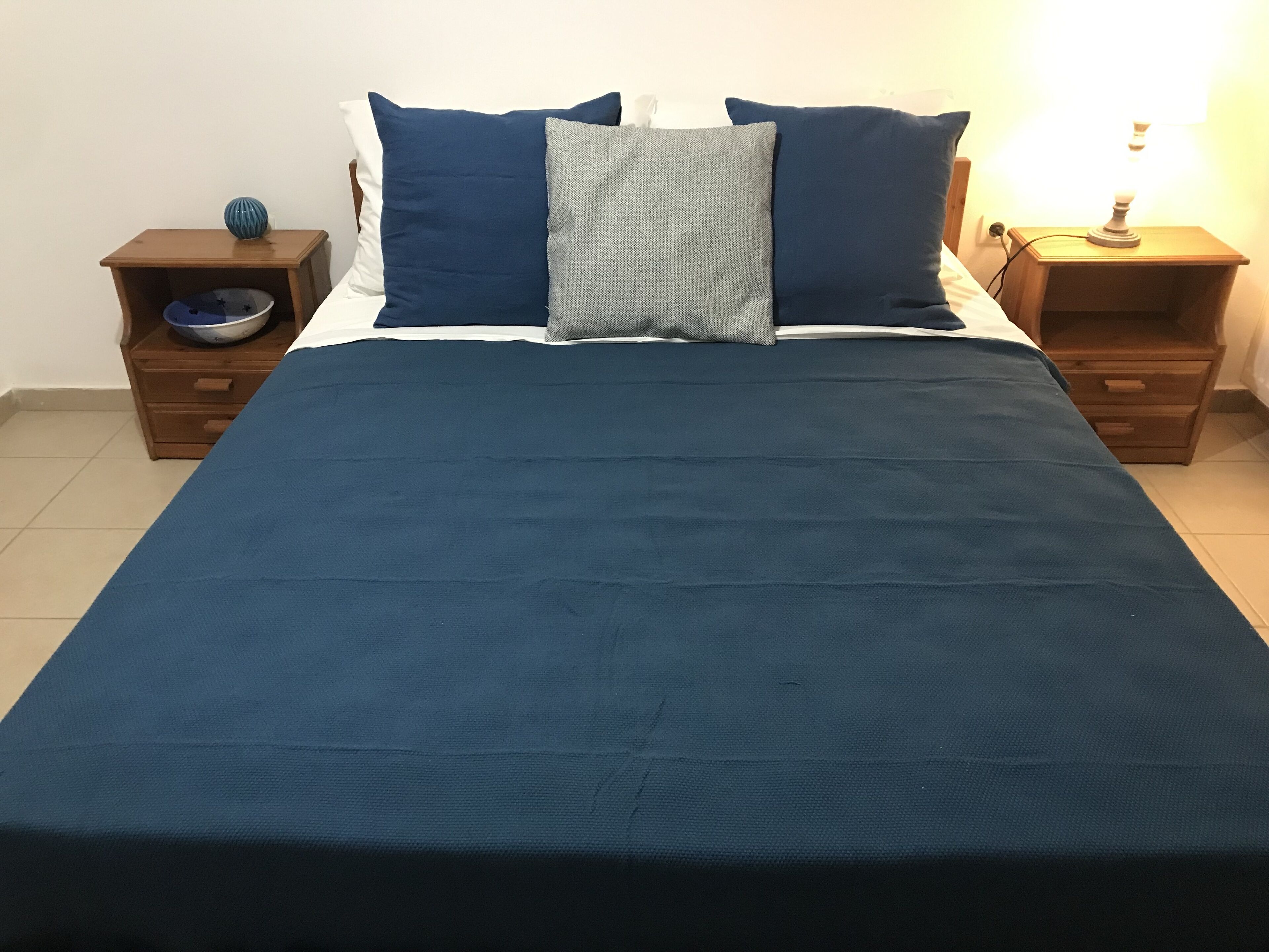 A bed with a blue bedspread, two pillows, and a bedside table with a lamp.