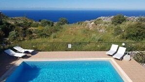Une piscine à l'eau d'un bleu cristallin, entourée d'une terrasse carrelée. Des chaises longues sont disposées le long du bord de la piscine, et on peut admirer la mer et les collines au loin.