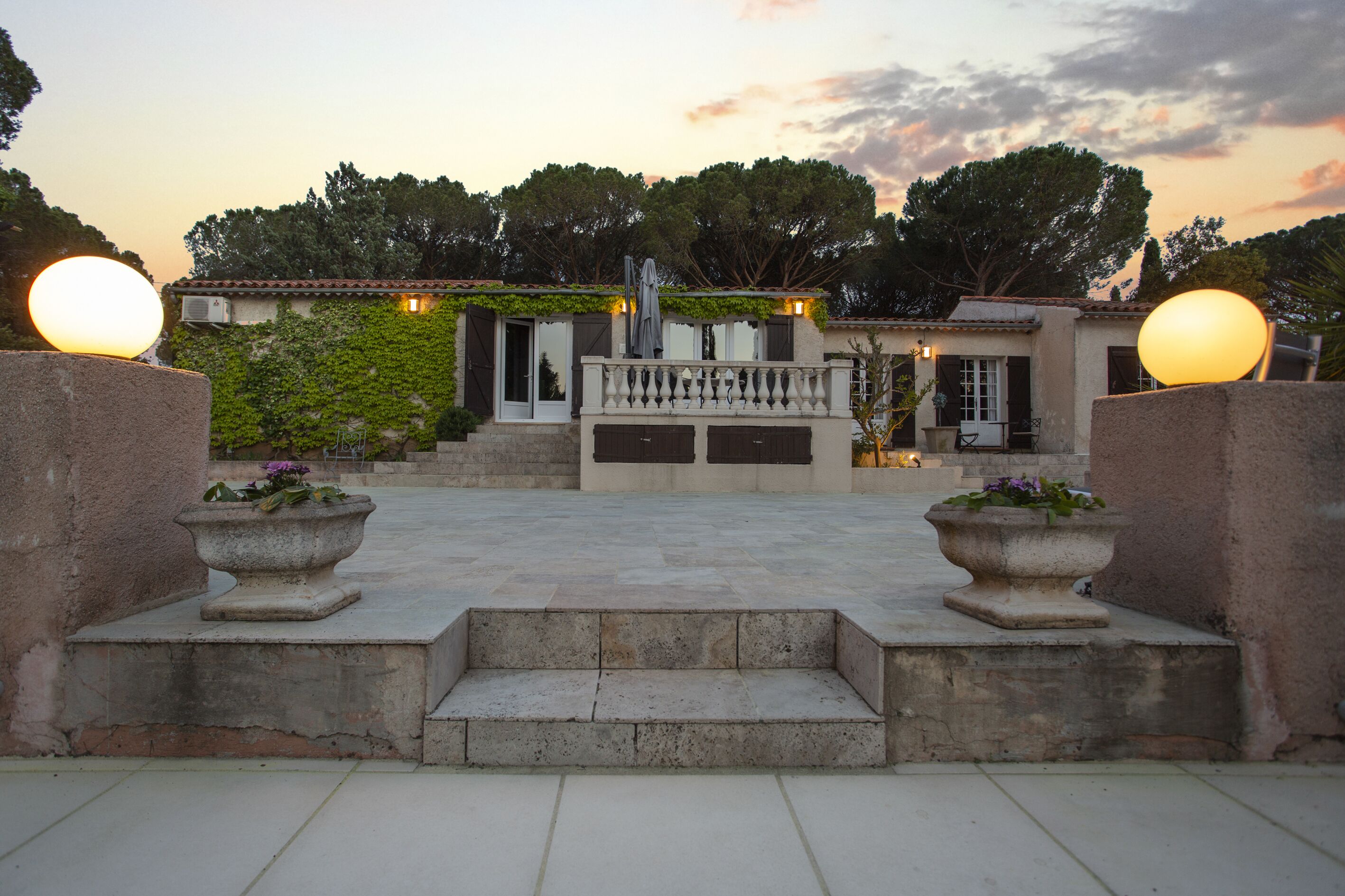 A courtyard with a stone staircase leading to a building with a balcony. There are two large spherical lights on either side of the entrance.