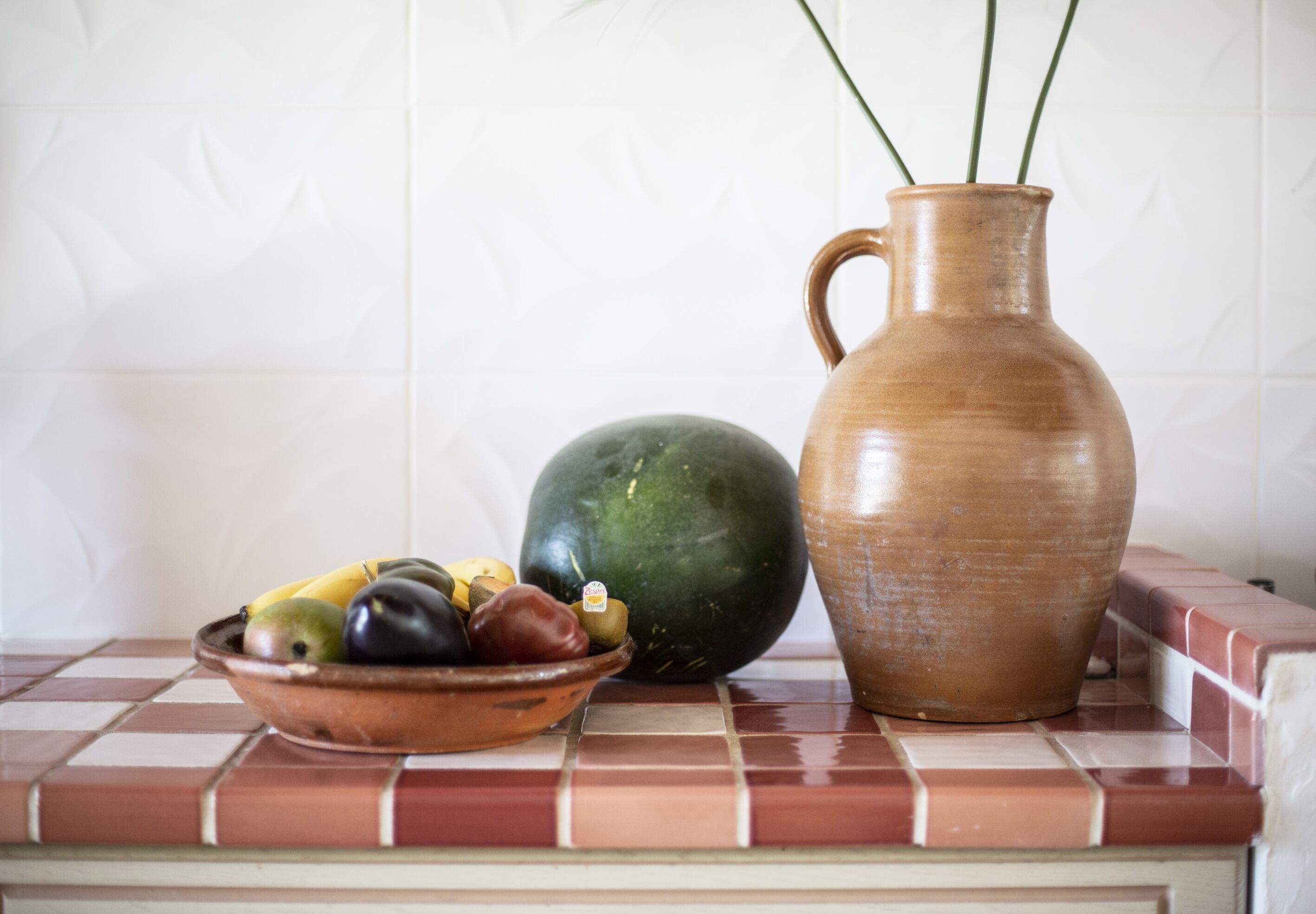 Una encimera de cocina con un cuenco de fruta, un melón grande y una jarra de cerámica.