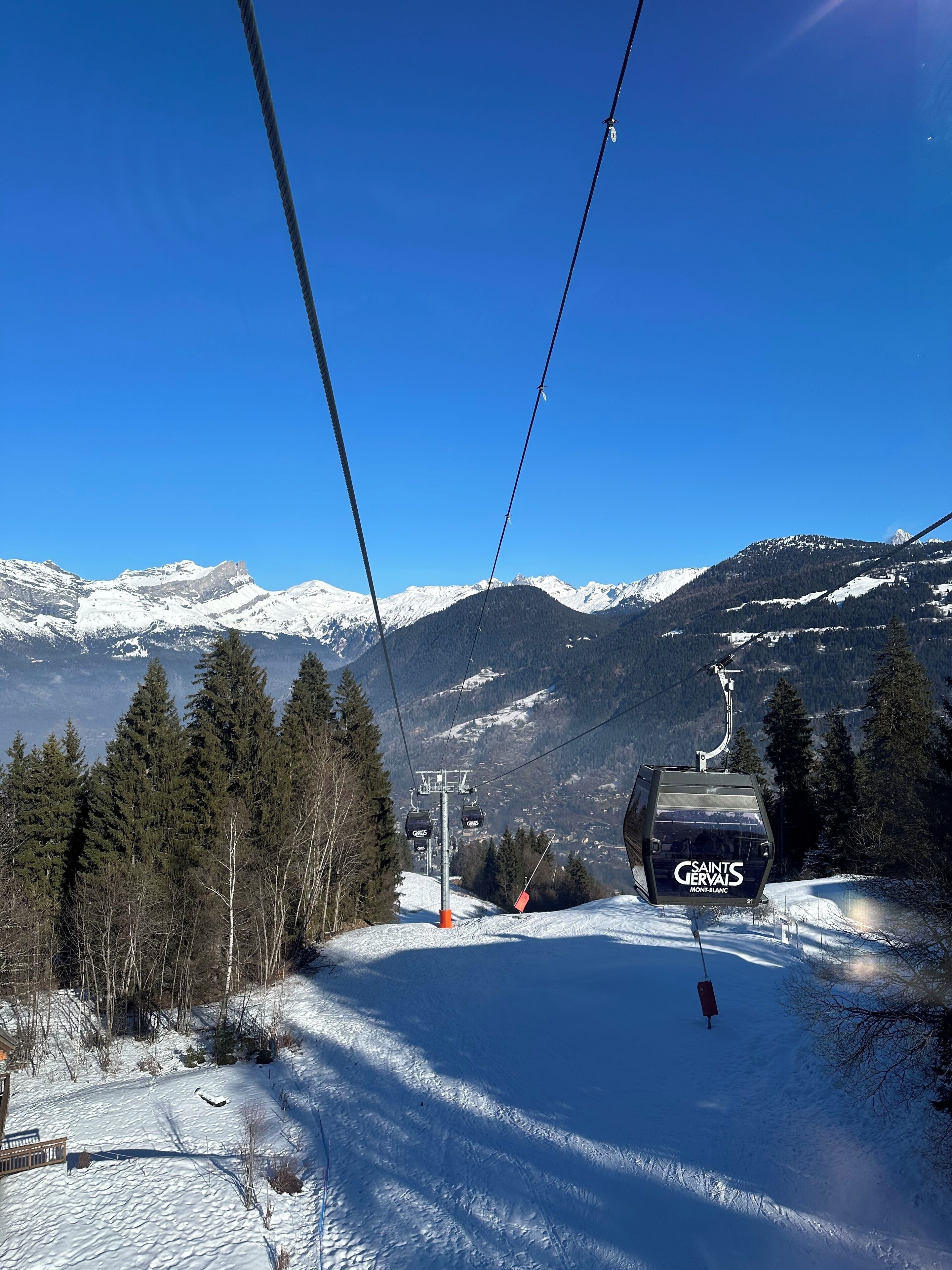 A ski lift chair with "SAINTS GERVAI" branding, a snowy mountain landscape, and a clear blue sky.