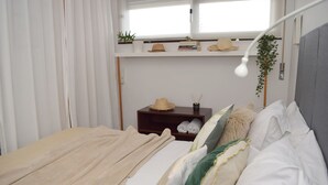Una cama bien hecha con una manta color beige, una mesita de noche con un sombrero y libros, y una planta colgante.