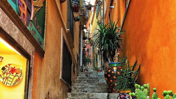 A narrow street with stone steps leading to a higher level, flanked by buildings with colorful facades and various potted plants.