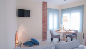 A modern living room with a white sofa, a round dining table with chairs, a TV mounted on the wall, and a brick column.