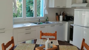 Eine Küche mit weißen Schränken, einem Holztisch mit Untersetzern, einem Kühlschrank und einem Fenster mit Vorhängen.