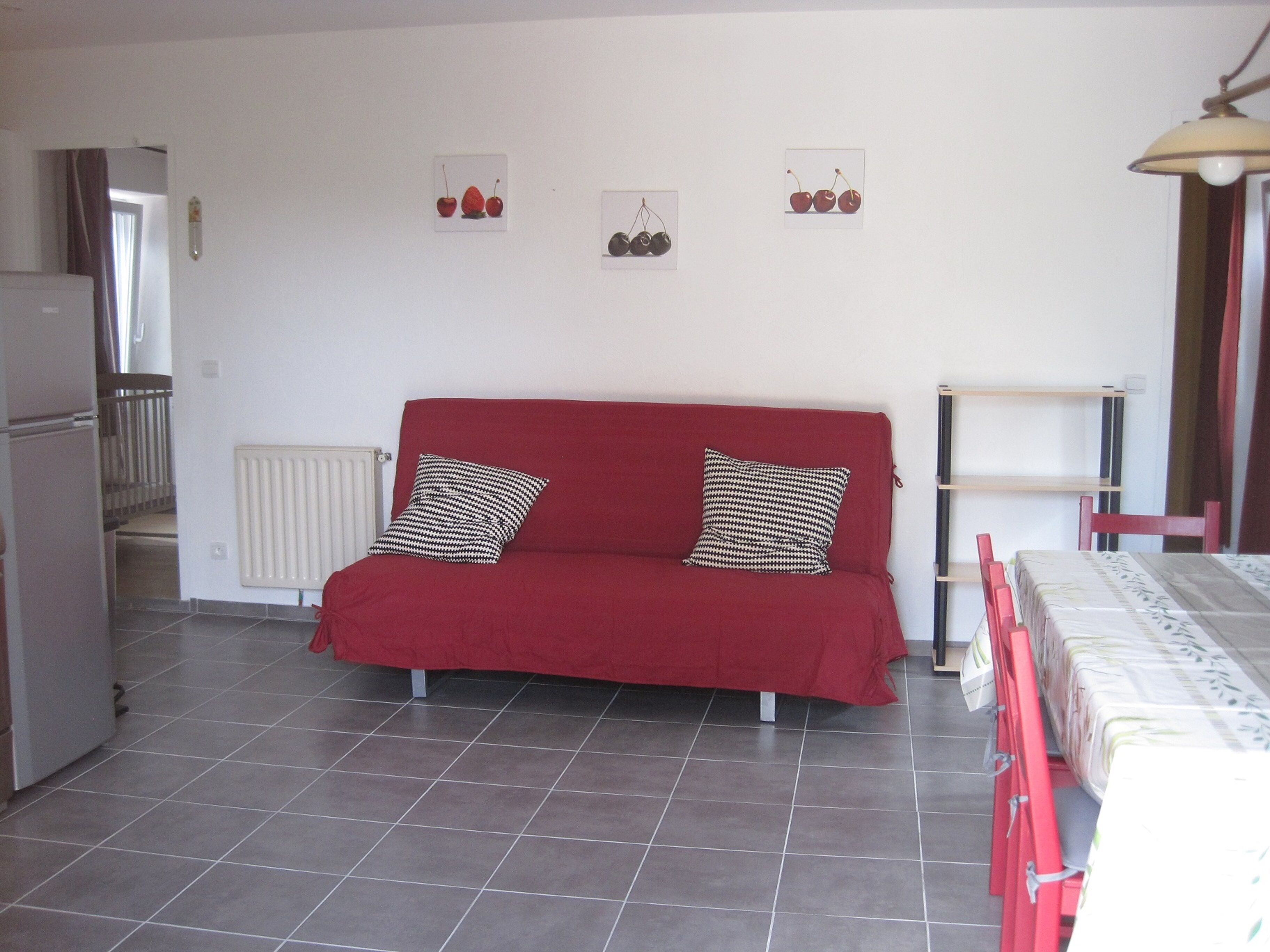 A red sofa with patterned pillows, a radiator, a small table with a lamp, and wall-mounted pictures of cherries.