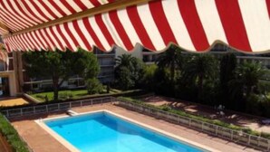 Une piscine surmontée d’un auvent à rayures rouges et blanches, entourée de verdure et avec un bâtiment visible au loin.