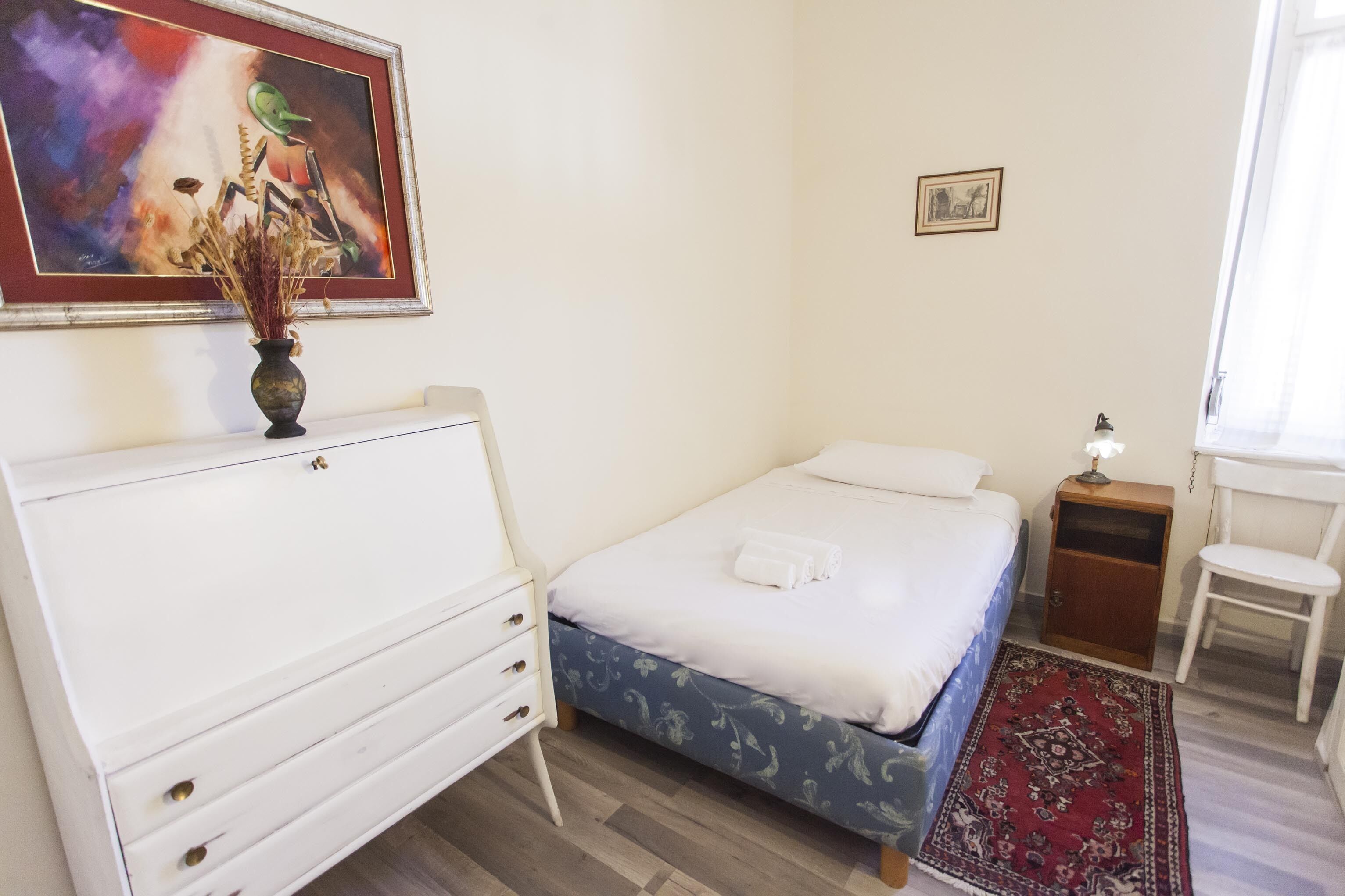 Ein Schlafzimmer mit einem Bett, einem Nachttisch, einem Sessel, einer Kommode, einem Gemälde und einem Teppich.