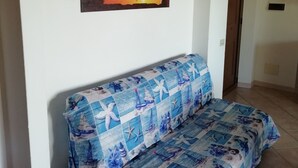 Una habitación con un sofá cubierto por una manta de diseño azul y blanco, un cuadro de un atardecer sobre el agua en la pared, y dos lámparas fijadas a la pared.