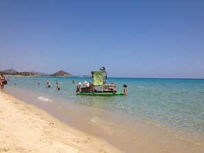 Ein Strand mit klarem türkisblauem Wasser, einer Rettungswache und Menschen, die schwimmen und am Ufer entlanggehen.