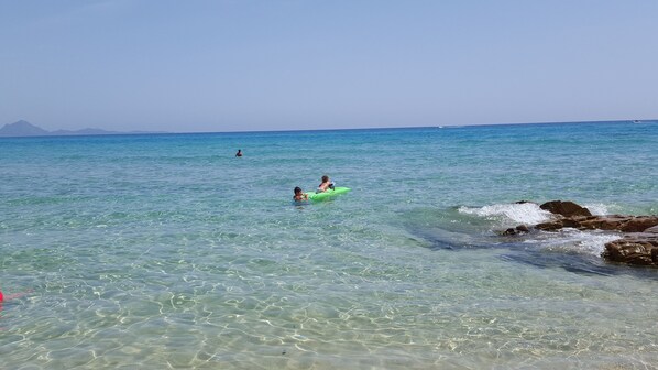 Ein sauberer Strand mit ruhigem Wasser, einer felsigen Küste und Bergen im Hintergrund.