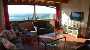 Un salón con un sofá, dos sillones, una mesa de centro de vidrio, un televisor sobre un mueble bajo y vistas del paisaje a través de amplios ventanales.