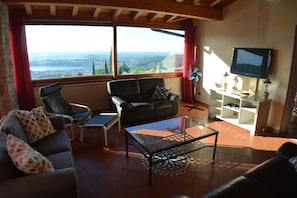 Un salón con un sofá, dos sillones, una mesa de centro de vidrio, un televisor sobre un mueble bajo y vistas del paisaje a través de amplios ventanales.