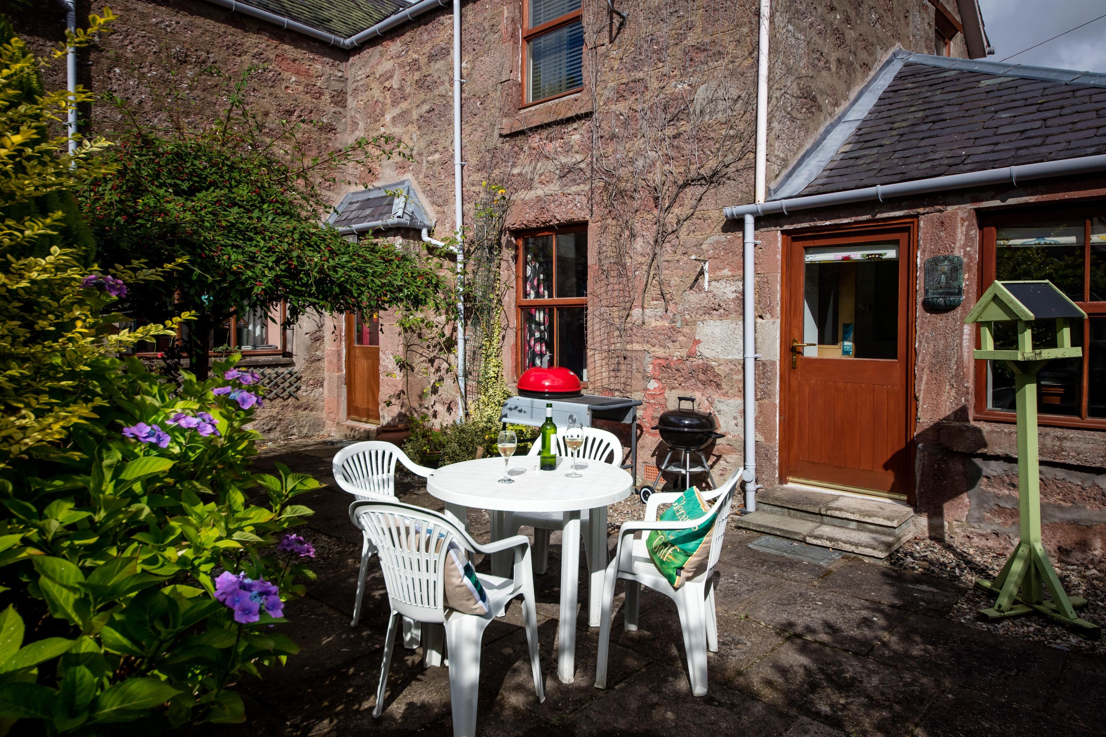 A brick building with a wooden door, a small garden with purple flowers, and a round table with chairs.
