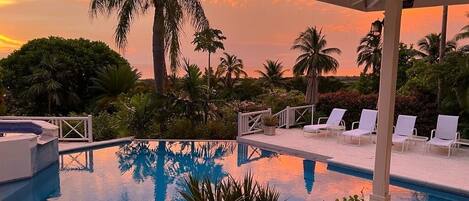 Zona de piscina con sillones de descanso y palmeras, reflejando un atardecer.