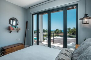 Un dormitorio con una cama grande, un banco de madera, un espejo redondo y vistas a un balcón con piscina y vegetación.