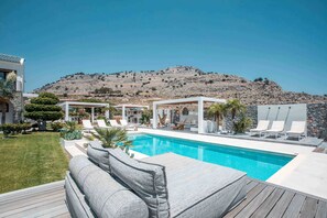 Zona de piscina con sillones de descanso, una terraza de madera y montañas al fondo.