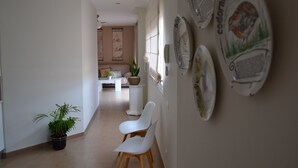 Een gang met een kamerplant, twee witte stoelen, een eethoek met een tafel en stoelen, en decoratieve borden aan de muur.