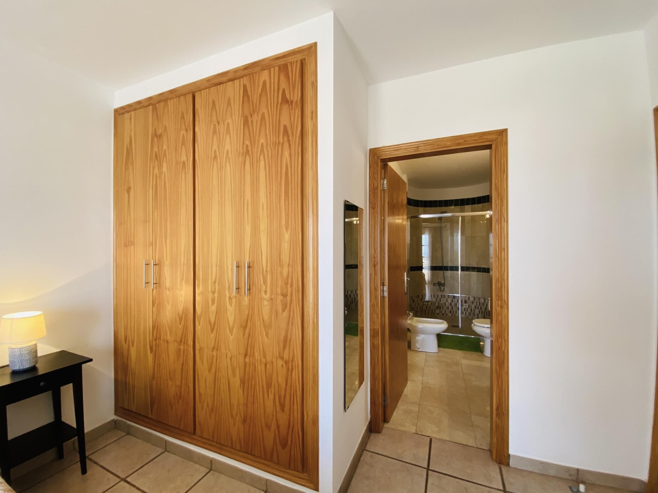 A room with a large wooden wardrobe, a small table with a lamp, and a bathroom visible through an open door.