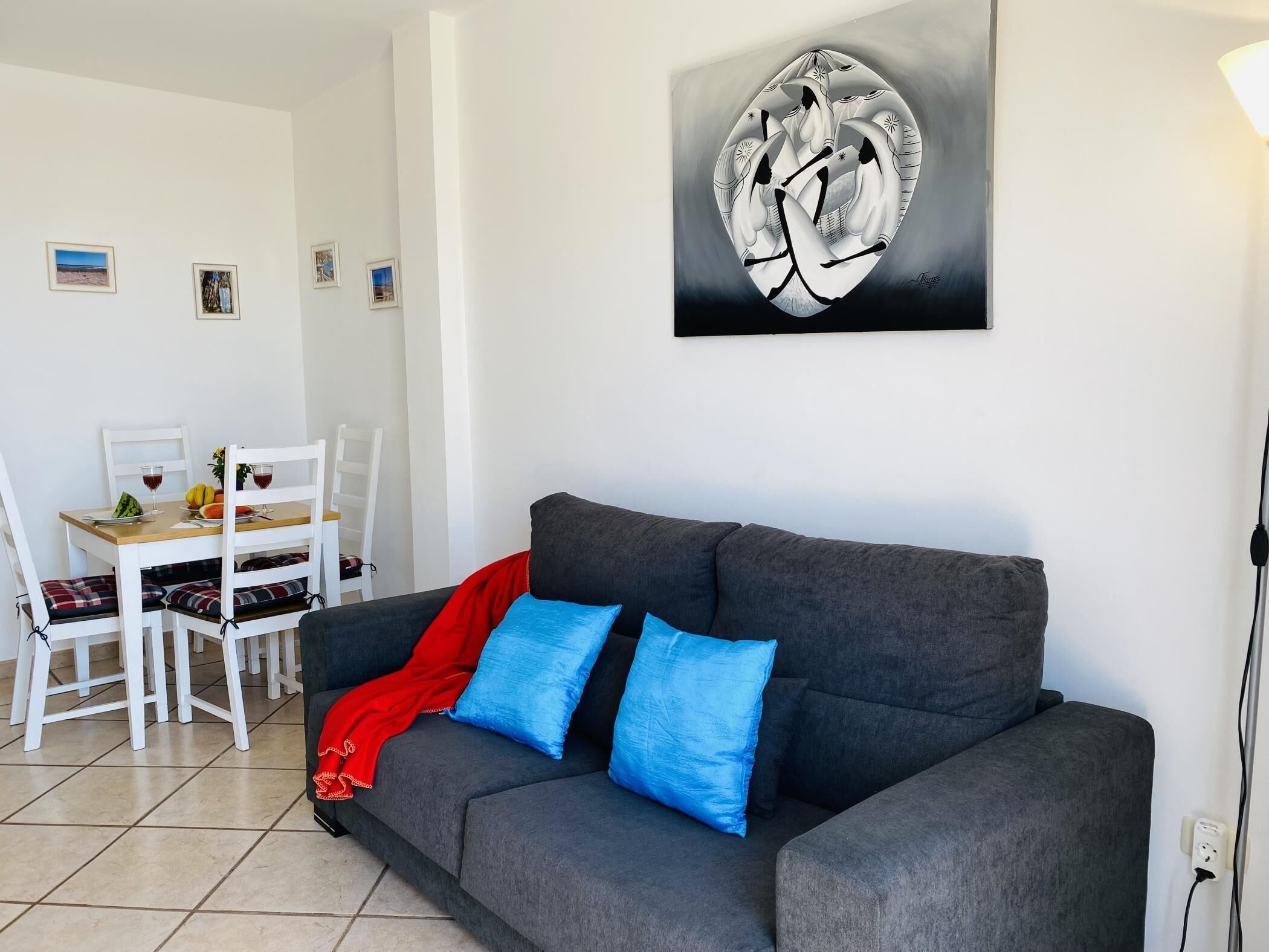 A living room with a grey sofa, two blue cushions, a red throw, and a dining area with a wooden table and white chairs.