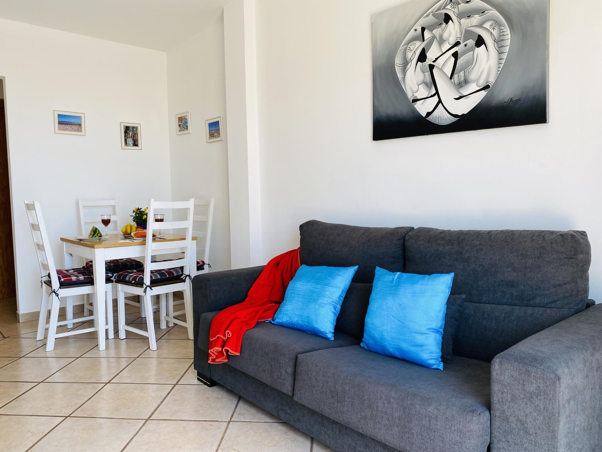 A living room with a grey sofa, two blue cushions, a red throw, and a dining area with white chairs and a table.