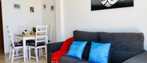 A living room with a grey sofa, two blue cushions, a red throw, and a dining area with white chairs and a table.