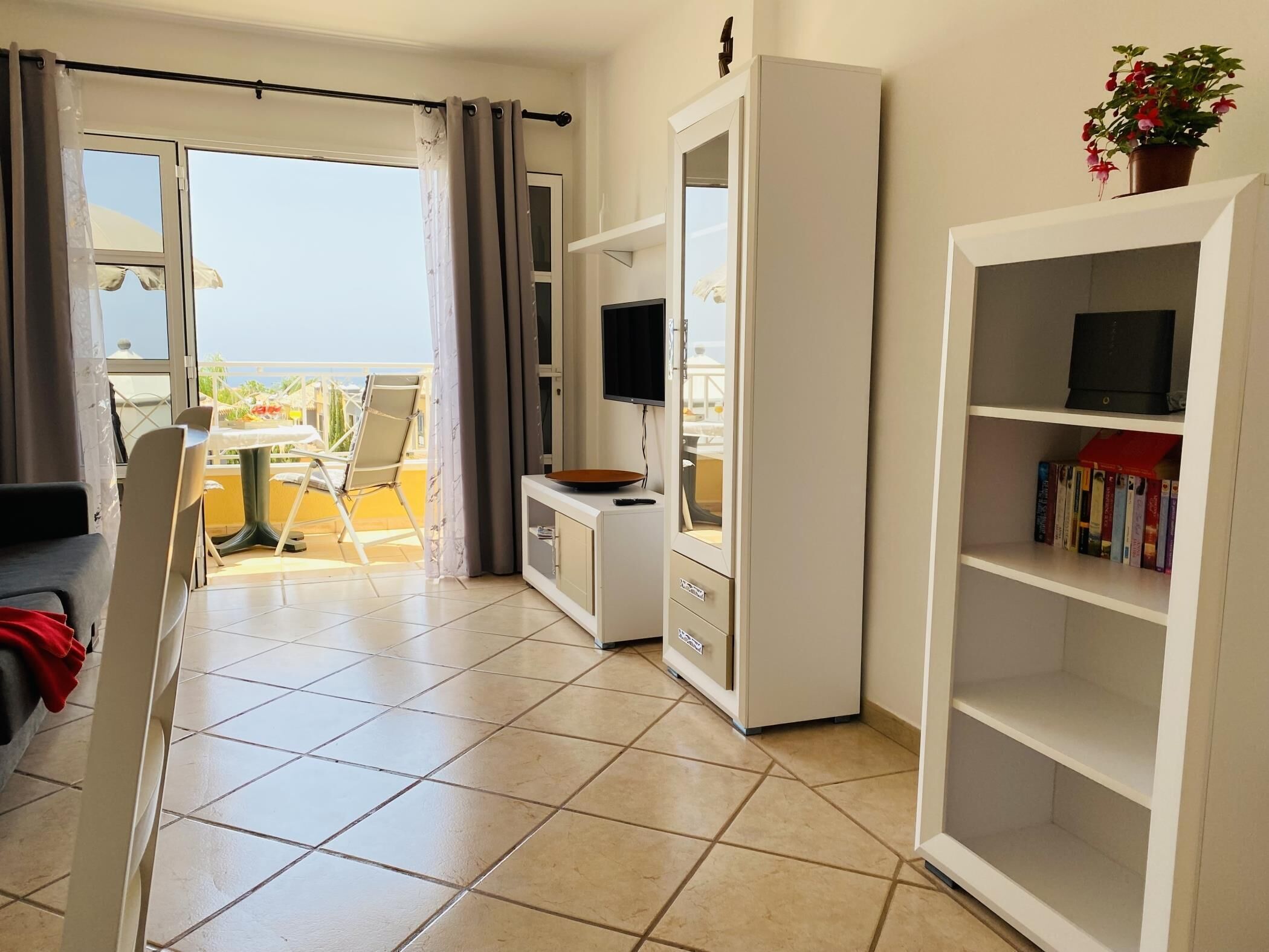 A living room with a sofa, a TV, a bookshelf, and a balcony with a table and chairs.
