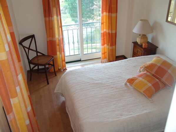 Un dormitorio con cama, una silla, una mesita de noche con lámpara y cortinas.
