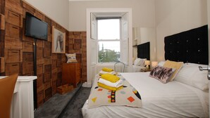 Une chambre d’hôtel avec un panneau mural en bois, un lit avec une literie blanche et un plaid coloré, un téléviseur à écran plat fixé au mur, et une fenêtre donnant sur la ville.