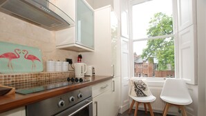 Une cuisine moderne équipée d’une cuisinière, d’un four et de meubles blancs. On y trouve deux chaises blanches près d’une fenêtre donnant sur des bâtiments à l’extérieur.