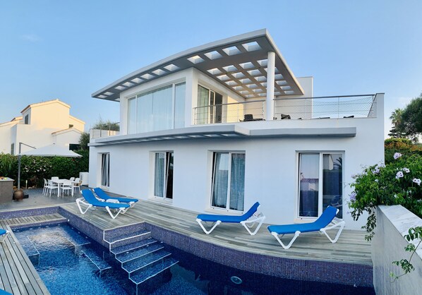 Una casa moderna de dos pisos con piscina, sillas de descanso azules y una pérgola.