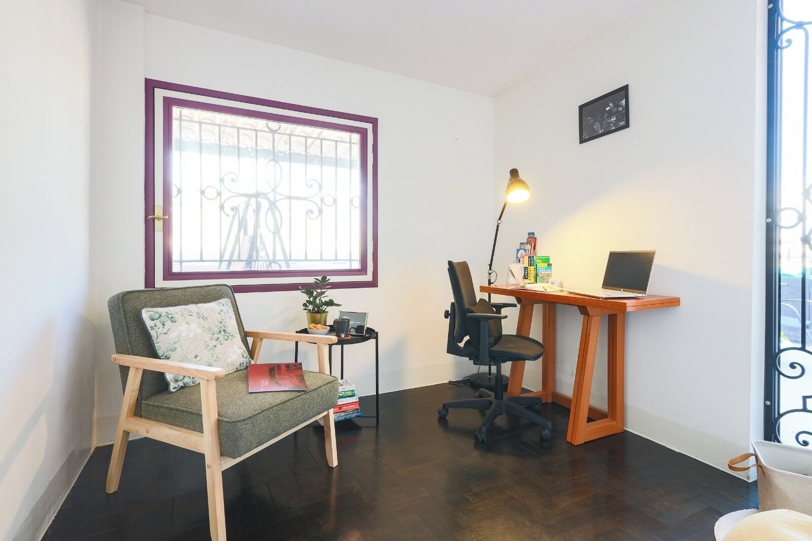 A small office with a wooden desk, a chair, a laptop, and a window with a metal grill.
