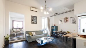 A modern living room with a sofa, coffee table, and dining area. There is a window with curtains, a plant, and artwork on the walls. - Penthouse with large terrace overlooking  Colosseum and  center of Rome. (Roma)
