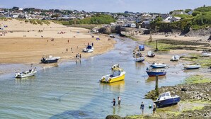 Un paese costiero con imbarcazioni ancorate in un canale stretto, una spiaggia sabbiosa e case sullo sfondo.
