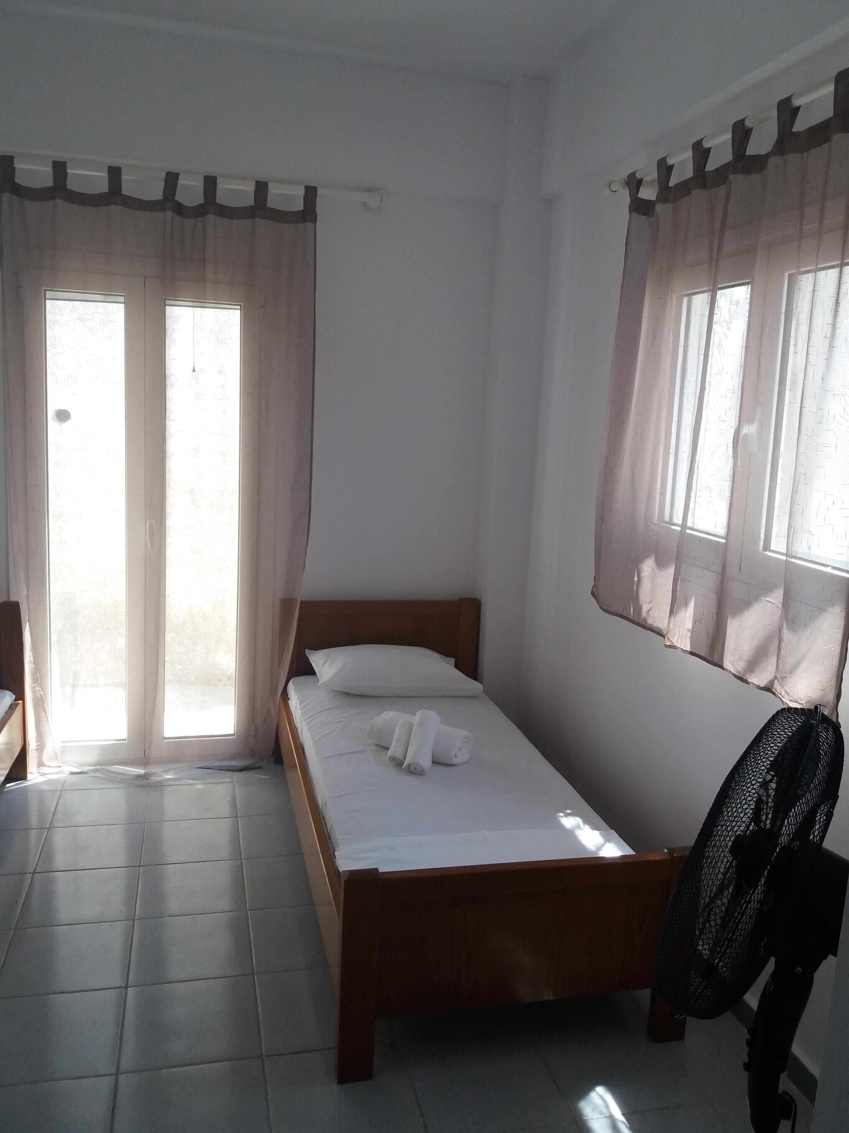 A single bed with a wooden frame, white bedding, and a black fan.