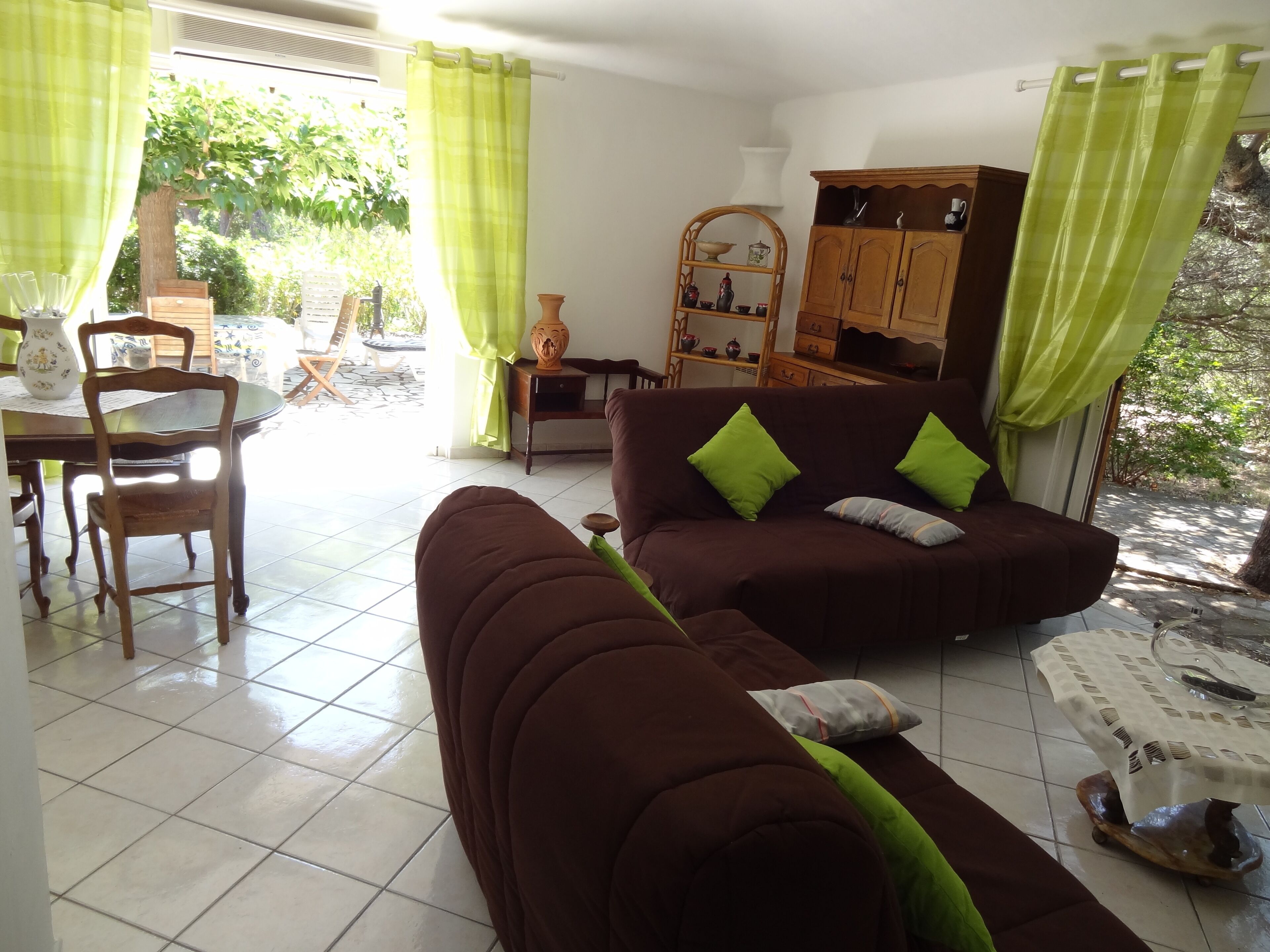 Un salon avec un canapé marron, des coussins verts, un meuble en bois et un coin repas comprenant une table et des chaises.