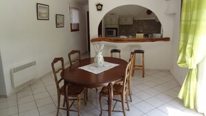 Ein Essbereich mit einem runden Holztisch, sechs Stühlen und einer Vase mit Gläsern. Im Hintergrund ist eine Küche durch einen Durchgang erkennbar.