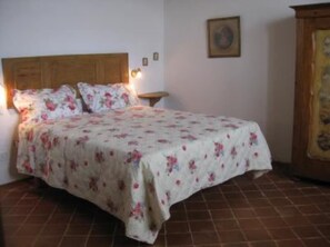 Un dormitorio con cabecera de madera, edredón estampado con motivos florales, una mesita de noche y un cuadro en la pared.