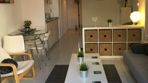 Ein modernes Wohnzimmer mit einem weißen Couchtisch, einer Couch und einem Essbereich mit einem Glastisch und Stühlen. Im Hintergrund ist eine Küche mit Schränken und einem Deckenventilator zu sehen.