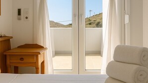 Une chambre avec un lit, une table de chevet en bois, une paire de rideaux blancs et une vue sur la plage, visible à travers une porte vitrée.