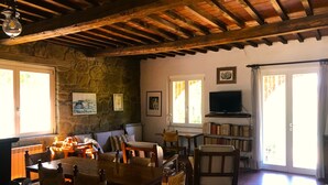 A rustic room with a stone wall, wooden ceiling beams, a dining table with chairs, a television, and a bookshelf. - House / Villa - Monte San Savino (Monte San Savino)