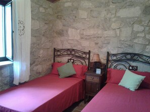 Une chambre avec deux lits, chacun agrémenté de draps et d’oreillers rouges, une table de chevet en bois avec une lampe, et un mur en pierre.