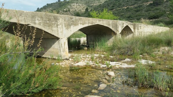 Un pont en béton enjambant un ruisseau aux eaux claires, avec une végétation luxuriante et des collines en arrière-plan.
