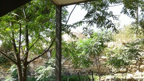 Un patio al aire libre con una mesa redonda, dos sillones a rayas, un barril rústico y un muro de piedra al fondo.