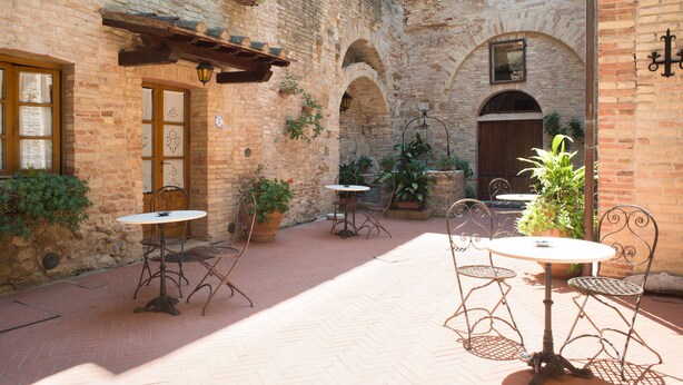 An outdoor courtyard with brick walls, arched doorways, and potted plants. There are metal tables and chairs arranged for outdoor dining. - San Gimignano Apartment in the Heart of the City for 3 People: Fast Wi-Fi, Concierge Service (San Gimignano)