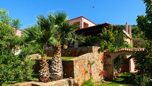 Una casa de piedra tradicional con techo de tejas rojas, rodeada de exuberante vegetación y palmeras.