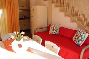 A room with a red sofa, white chairs, a small table with a vase of flowers, a kitchen area, and a staircase.