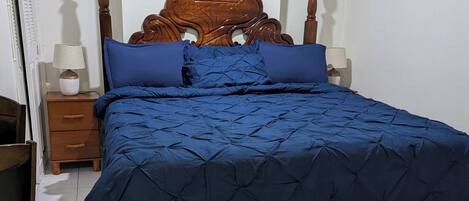 Een slaapkamer met een houten hoofdbord, een blauw beddegoed, twee nachtkastjes met lampen en een betegelde vloer.